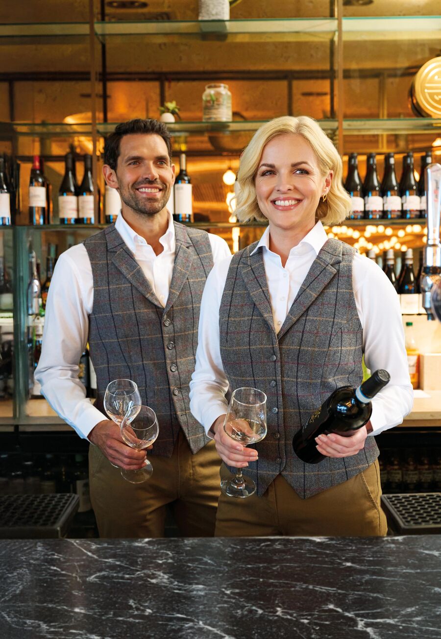 Two members of staff in grey and brown check tweed waistcoats behind a bar